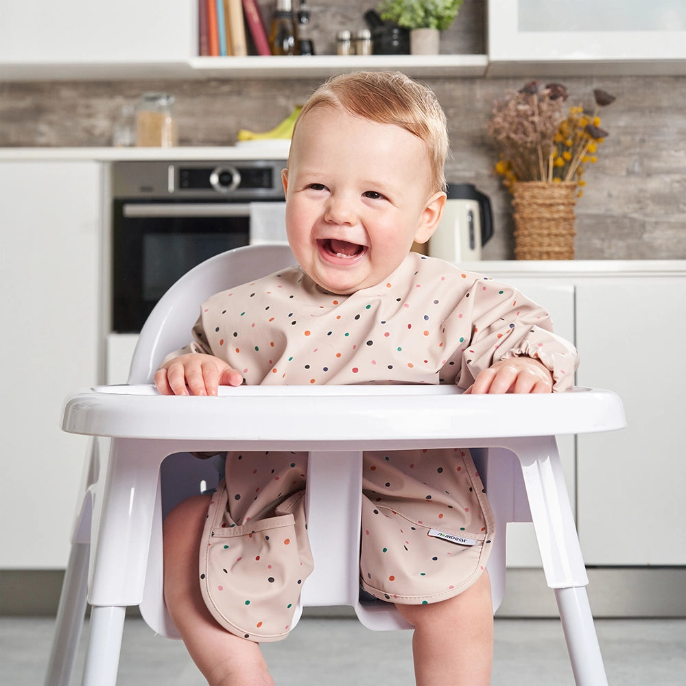 A Happy Smiling Baby, Sitting in a High Chair and Wearing a Confetti Pink Coverall Bib by Nombear